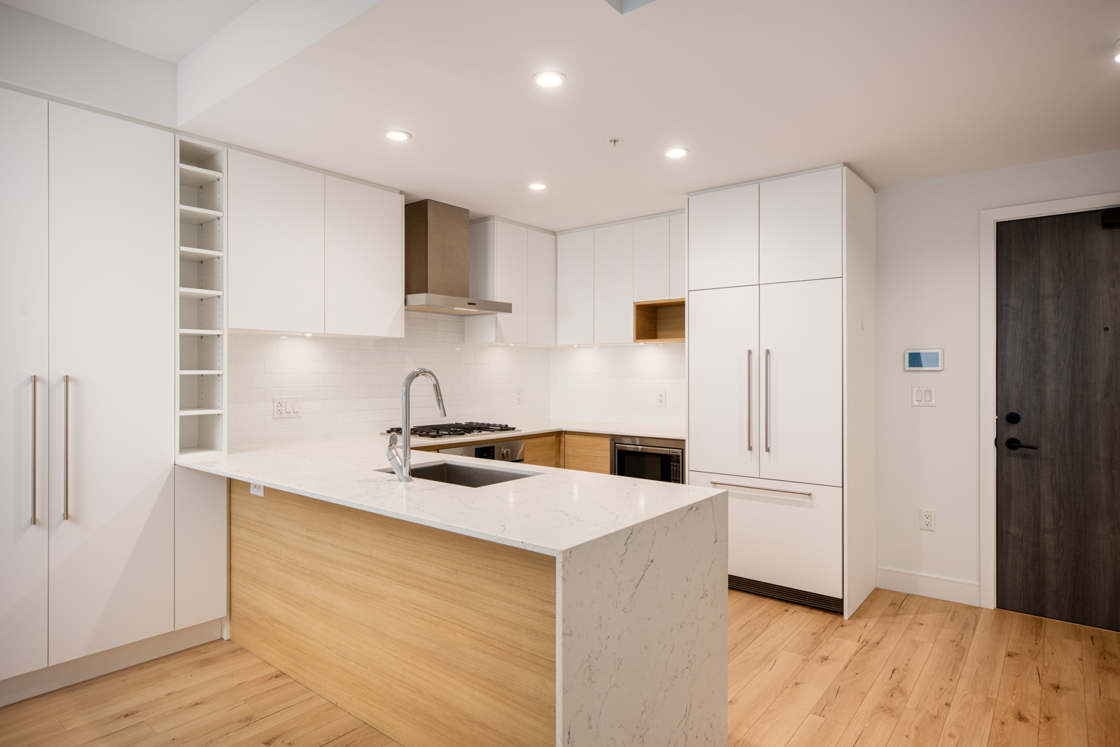 Modern kitchen with white cabinets, wood accents, stainless steel appliances, marble island with sink, and light wood flooring under recessed lighting.
