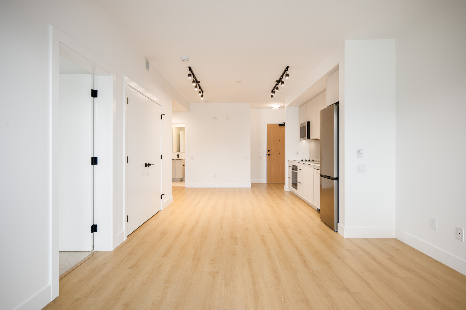 Modern, unfurnished apartment with light wood flooring, white walls, a kitchen area with stainless steel appliances, and track lighting on the ceiling. Multiple doors lead to other rooms.