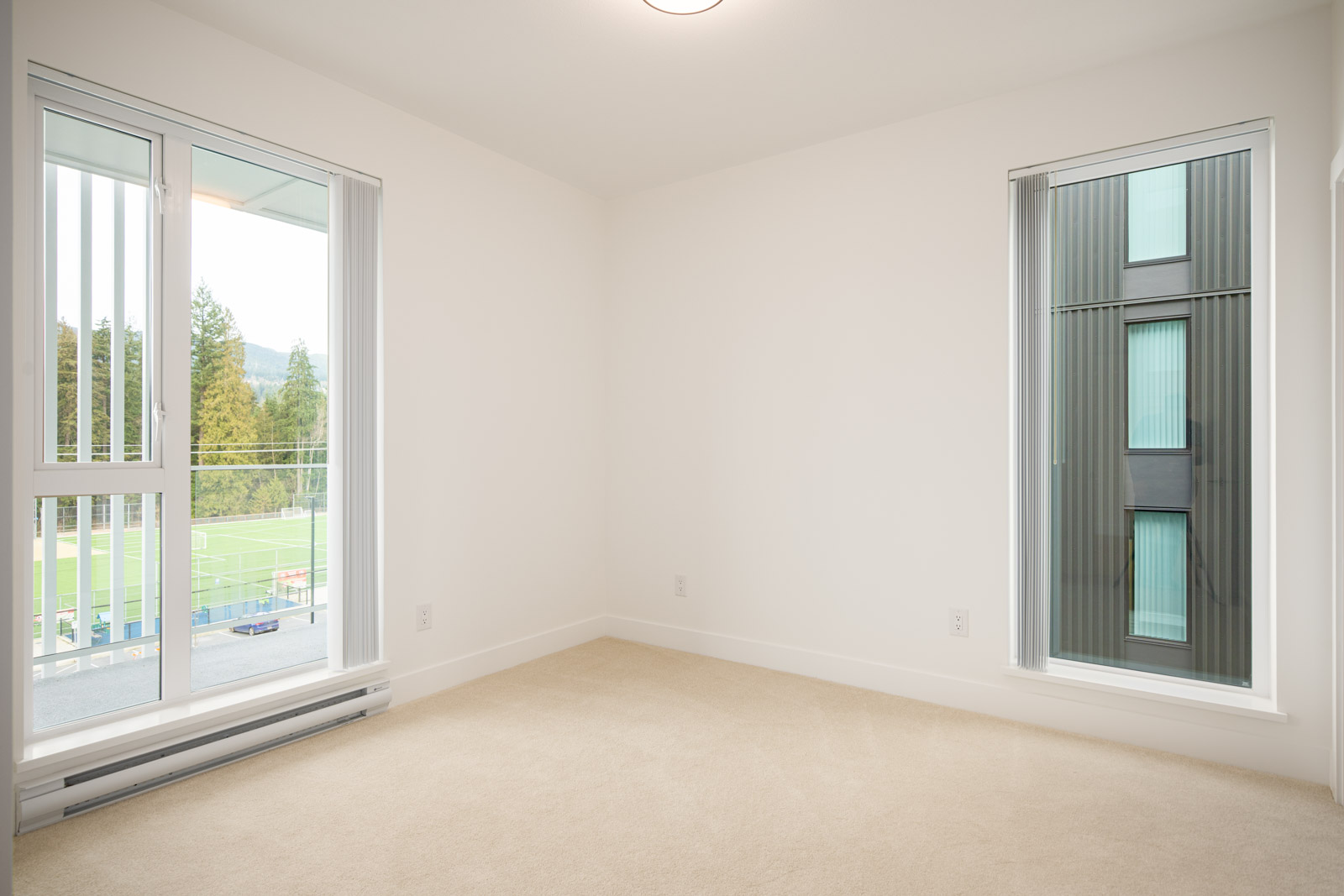 Empty room with beige carpet, white walls, and two large windows; one window overlooks a sports field and the other faces a dark exterior building wall.