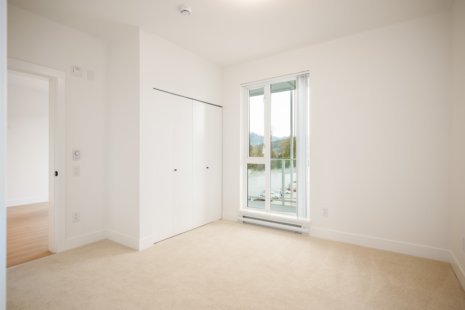 Empty bedroom with beige carpet, white walls, a closed closet, and a large window letting in natural light. A partially open door leads to another room.