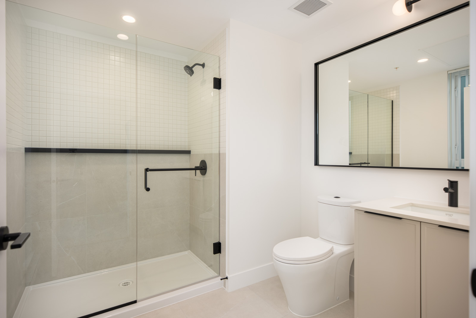 Modern bathroom with glass-enclosed shower, white toilet, beige vanity with sink, black fixtures, large mirror, and white tiled walls.