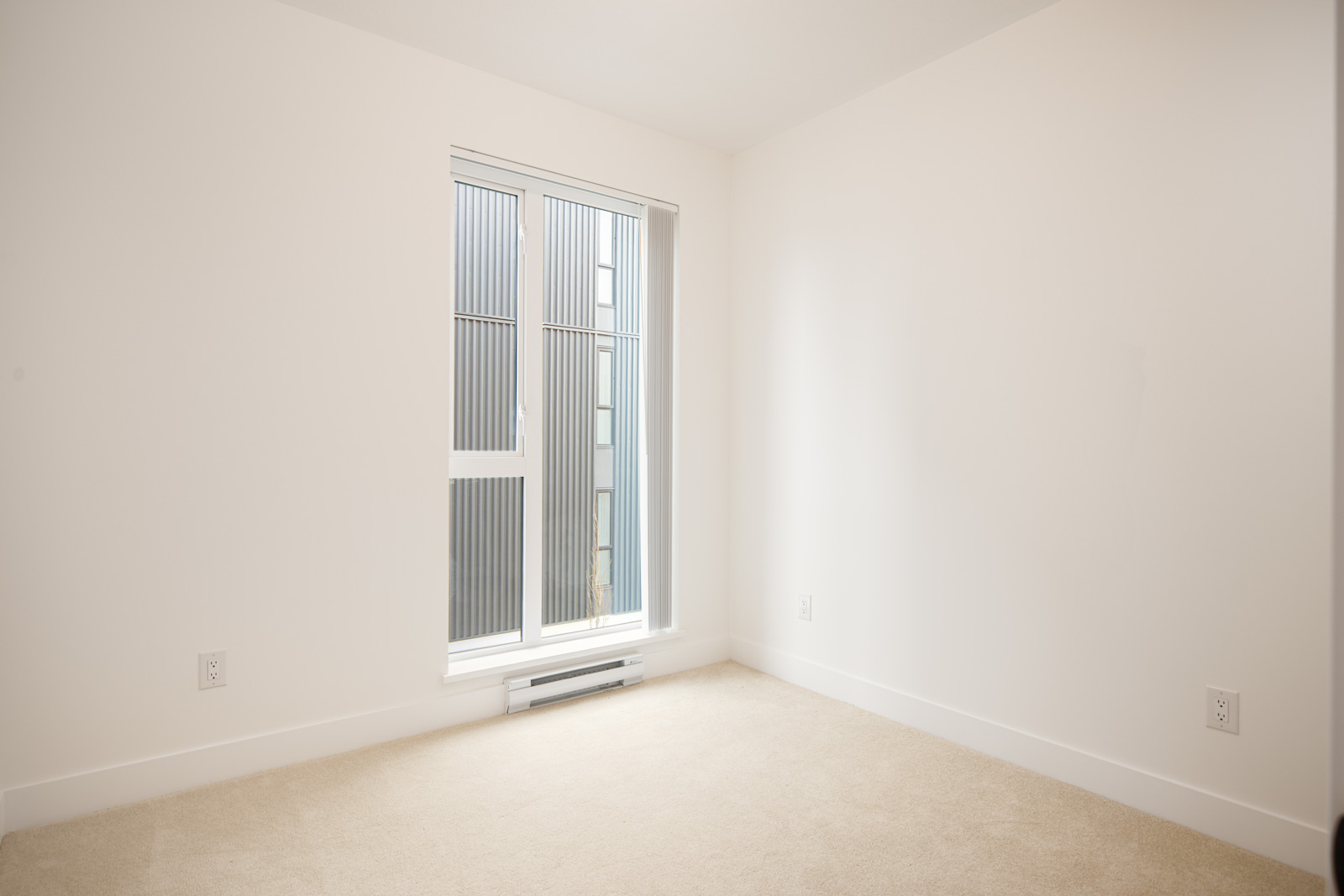 An empty room with beige carpet, white walls, and a large window with vertical blinds letting in natural light.