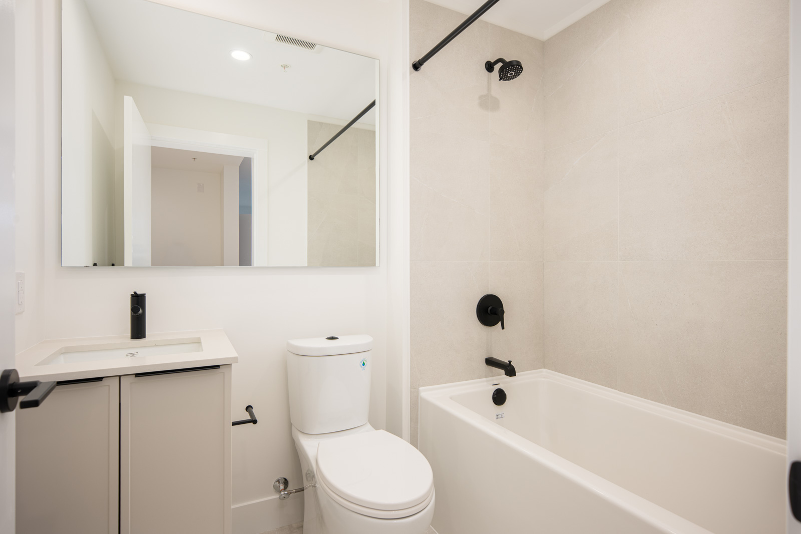 Modern bathroom with white walls, a bathtub-shower combo, toilet, vanity with sink, large mirror, and black fixtures.