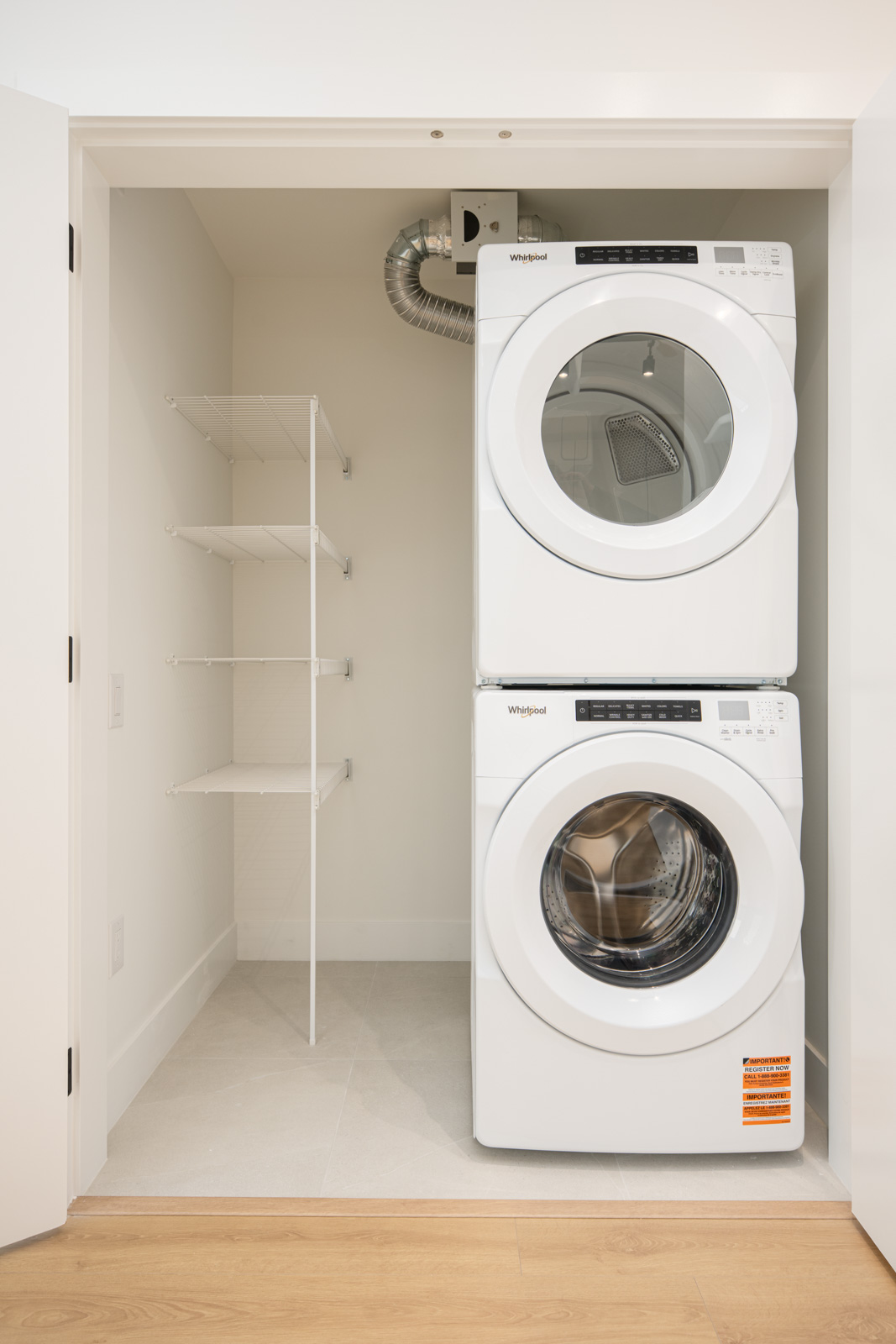 A small laundry closet with a stacked white washer and dryer on the right and five white wire shelves on the left.