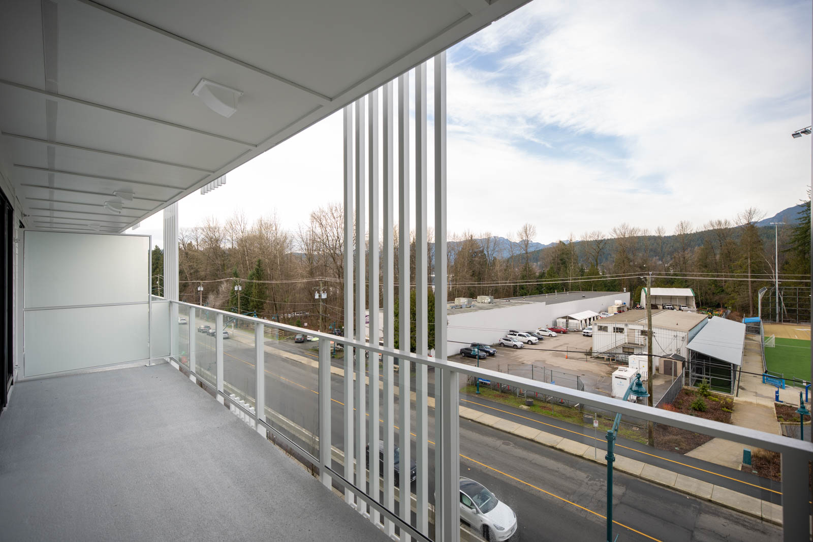 A view from a modern balcony overlooking a street, parking lot, fenced sports field, and trees with mountains in the distance under a partly cloudy sky.
