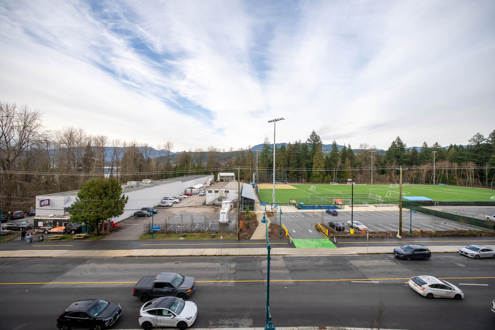 Wide view of a sports complex with green fields, parking lots, and adjacent parked cars, seen across a busy street under a cloudy sky.