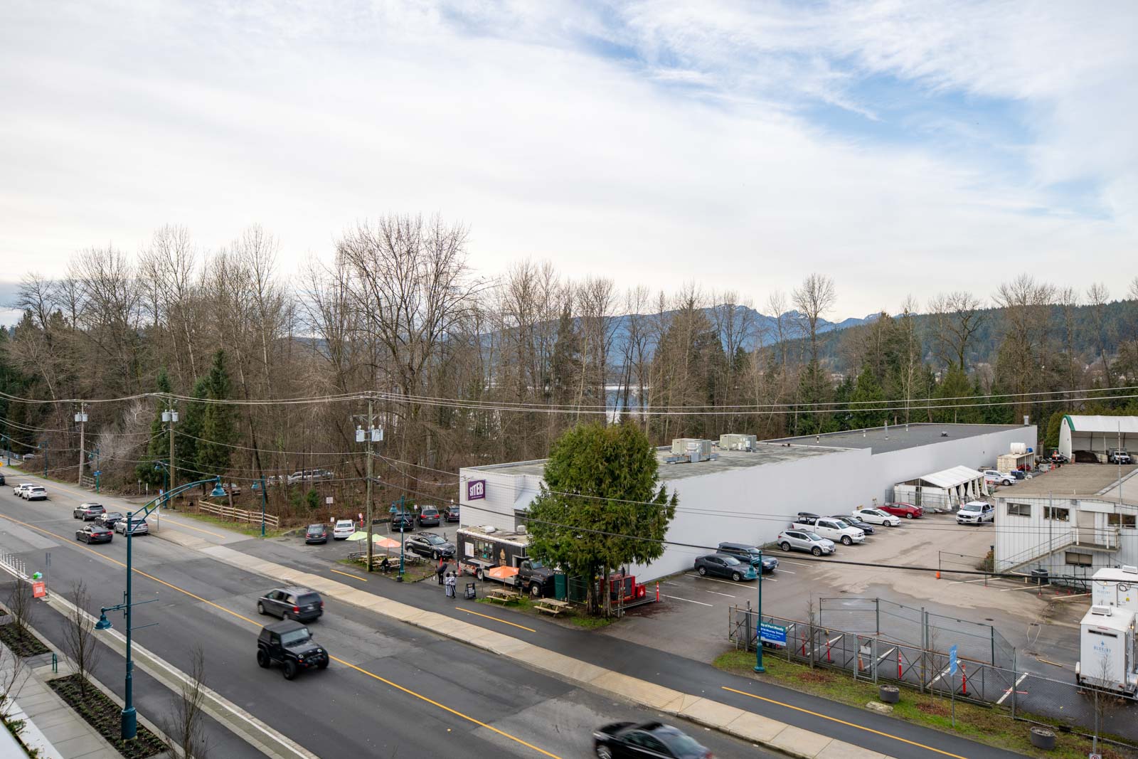 A wide view of a commercial building and parking lot beside a road with several cars, trees, and mountains visible in the background under a partly cloudy sky.