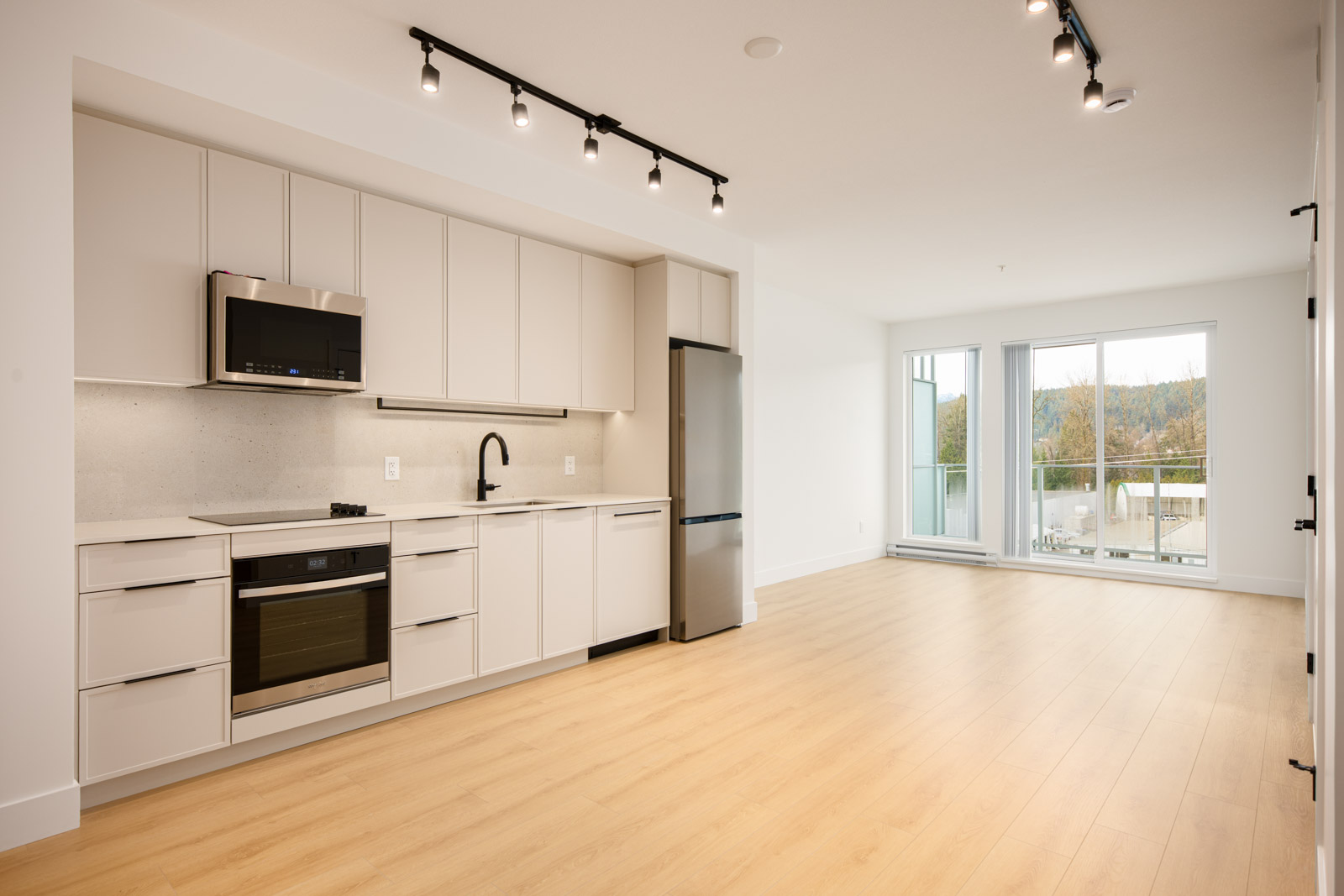 Modern apartment interior with light wood flooring, white cabinetry kitchen, stainless steel appliances, and large windows letting in natural light.