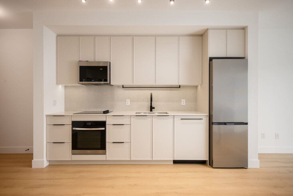 Modern minimalist kitchen with light cabinetry, stainless steel appliances, an electric stove, built-in microwave, single sink, and a refrigerator, set against a white wall and wood flooring.