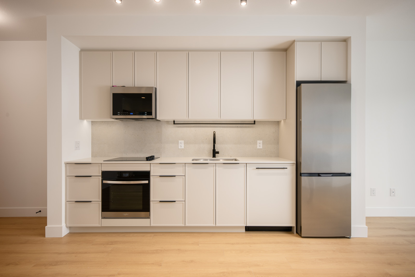 Modern minimalist kitchen with light cabinetry, stainless steel appliances, an electric stove, built-in microwave, single sink, and a refrigerator, set against a white wall and wood flooring.