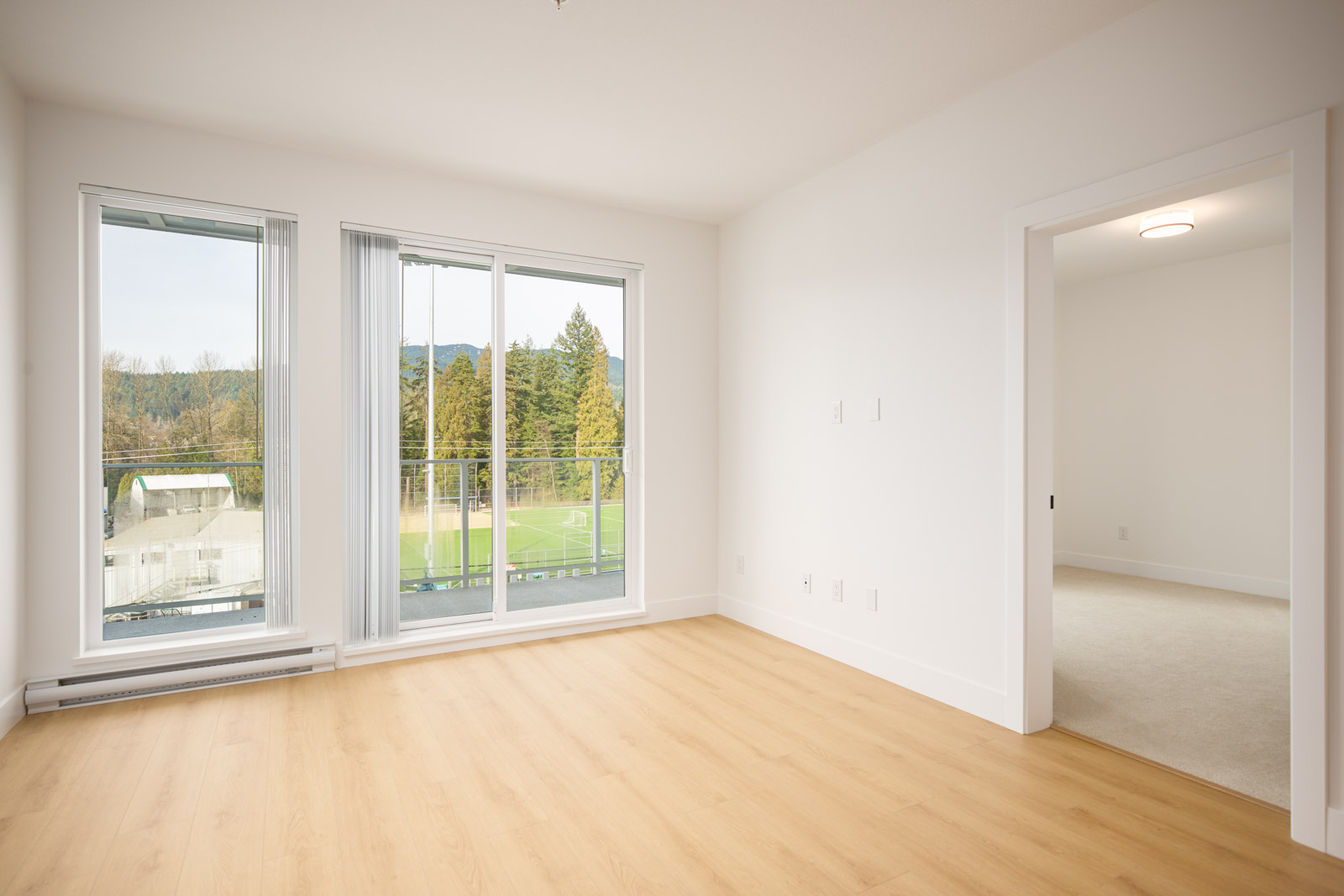 Empty, unfurnished room with light wood flooring, large windows and glass door leading to a balcony overlooking a soccer field, with an open doorway to an adjacent carpeted room.