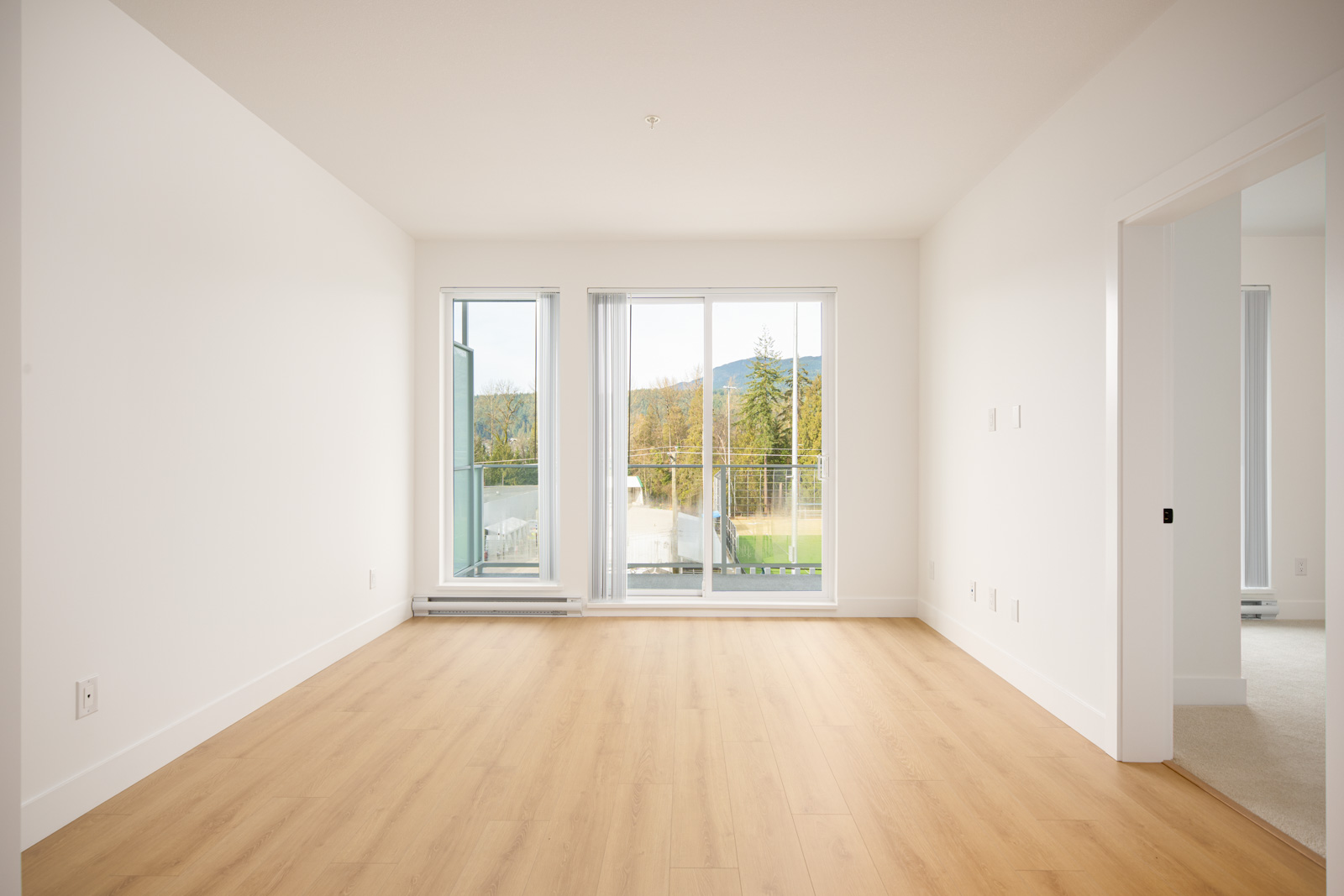 Empty, unfurnished room with light wood flooring, white walls, large windows, and sliding glass doors opening to a balcony with an outdoor view. Doorway leads to another room.