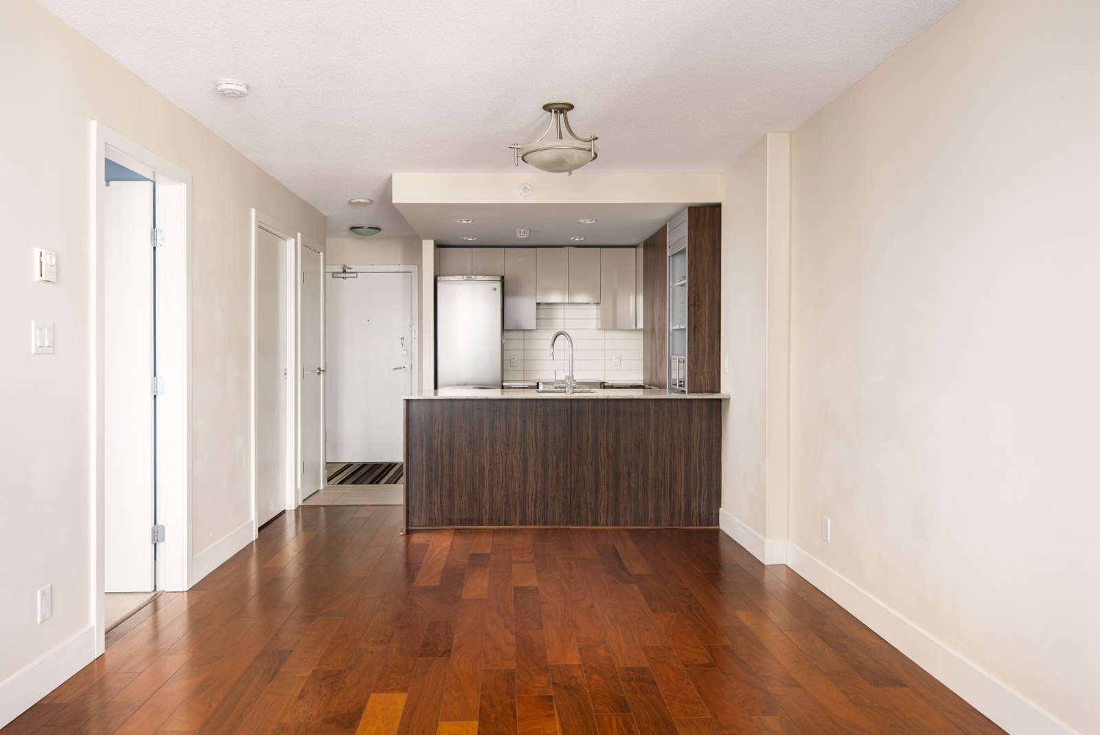 A modern, unfurnished apartment interior with wood floors, a kitchen featuring white cabinetry and stainless steel appliances, and neutral-colored walls.