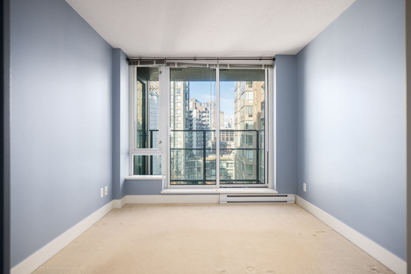 Empty room with light blue walls, beige carpet, and large floor-to-ceiling window leading to a balcony with a view of city buildings.