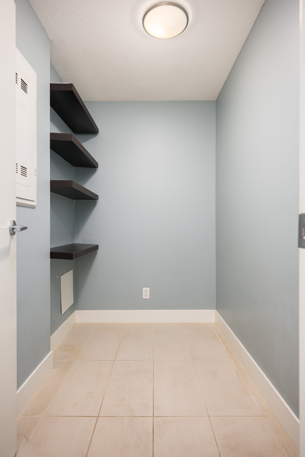 Small empty room with light blue walls, beige tile flooring, a ceiling light, open door, wall shelves, electrical panel, and wall vent.