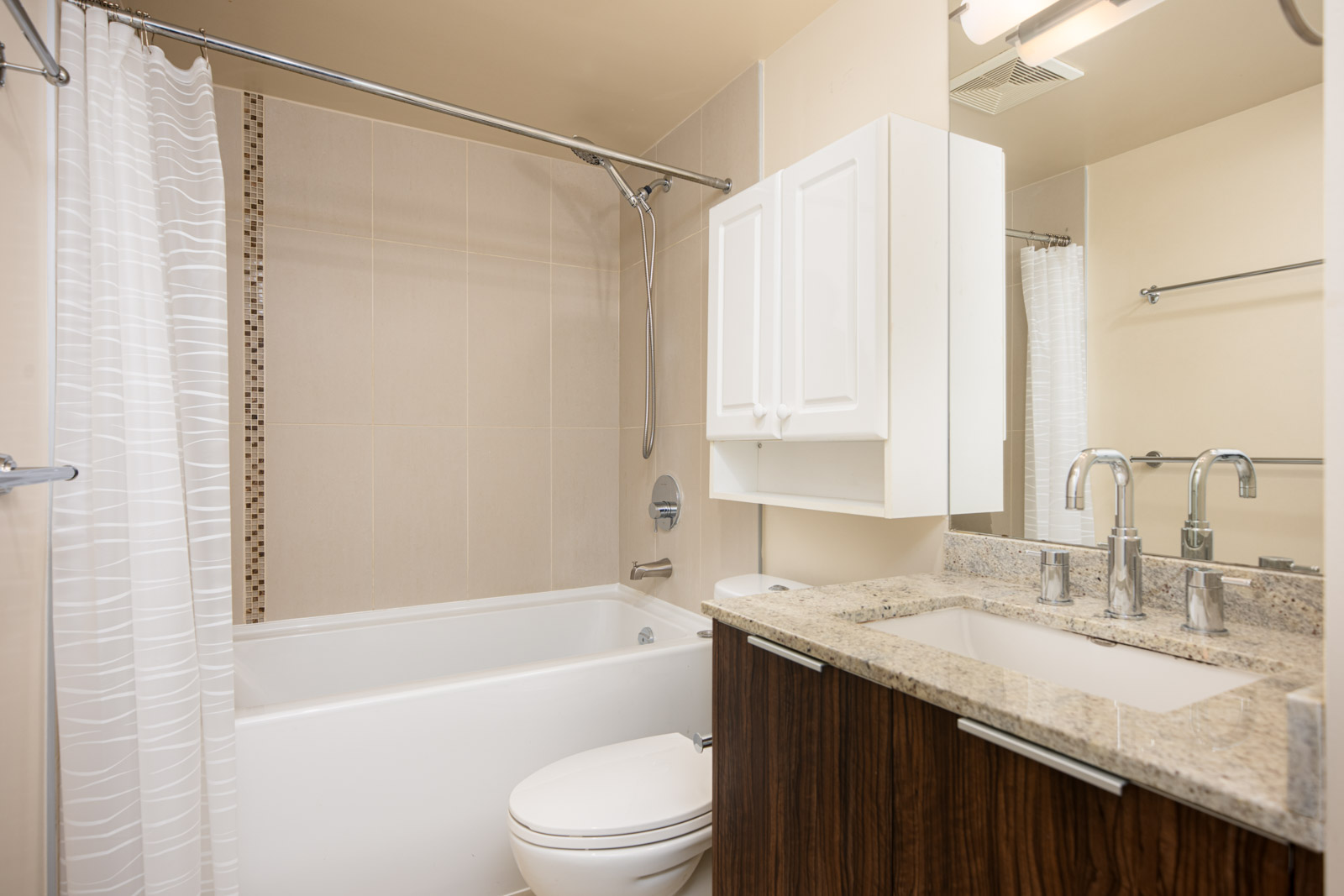 Modern bathroom with a white bathtub and shower, toilet, granite countertop with double sink, wall-mounted cabinet, and a large mirror.