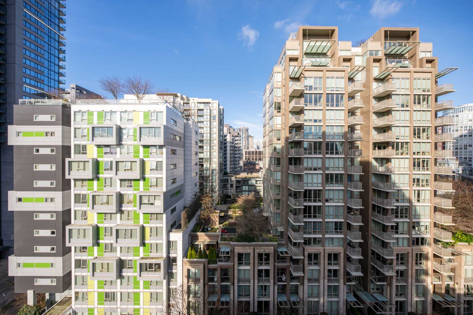 Modern high-rise apartment buildings with large windows and balconies in an urban area, separated by a landscaped courtyard, under a clear blue sky.