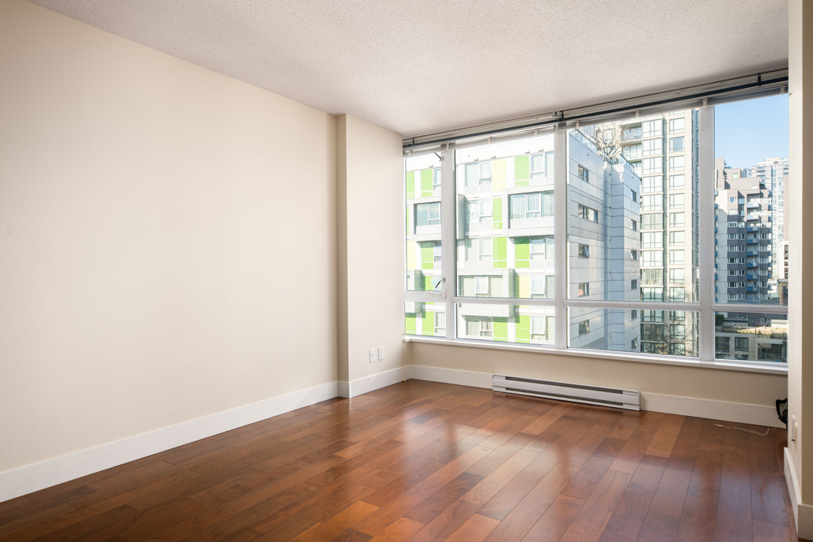 Empty room with hardwood floors, beige walls, large windows, and a view of nearby modern apartment buildings.