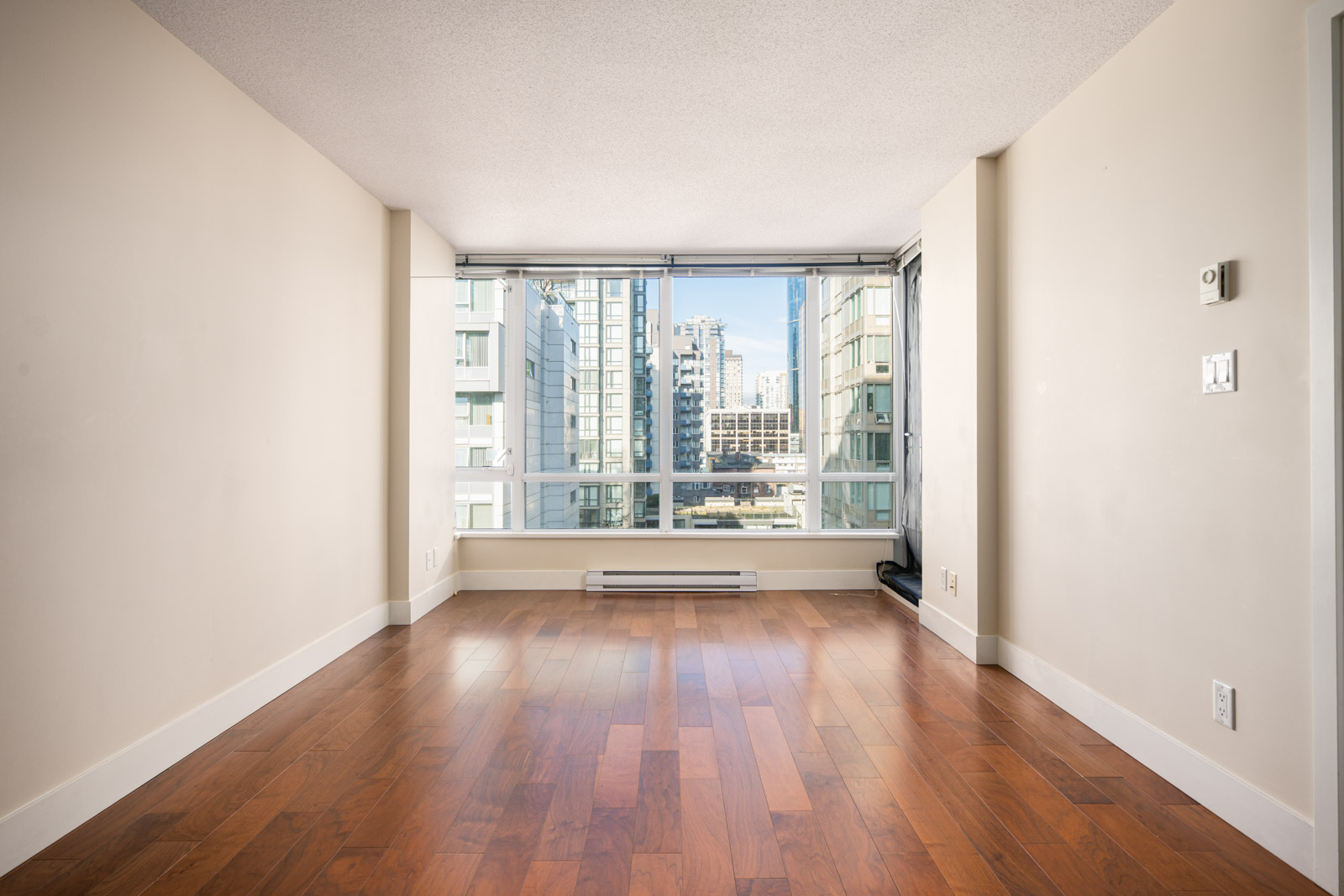 Empty room with light beige walls, wood flooring, and large floor-to-ceiling windows showing a cityscape outside.