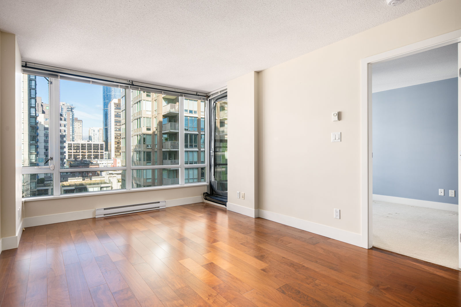 Unfurnished apartment room with large windows, wood flooring, and cityscape view; adjoining carpeted bedroom visible through open doorway.