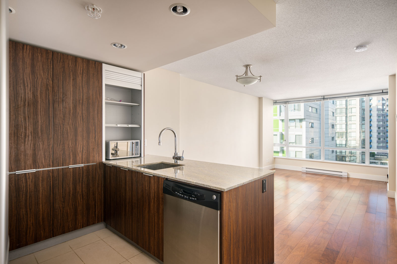 Modern apartment kitchen with wood cabinets, stainless steel appliances, granite countertop, and open plan leading to a bright living area with large windows and hardwood floors.