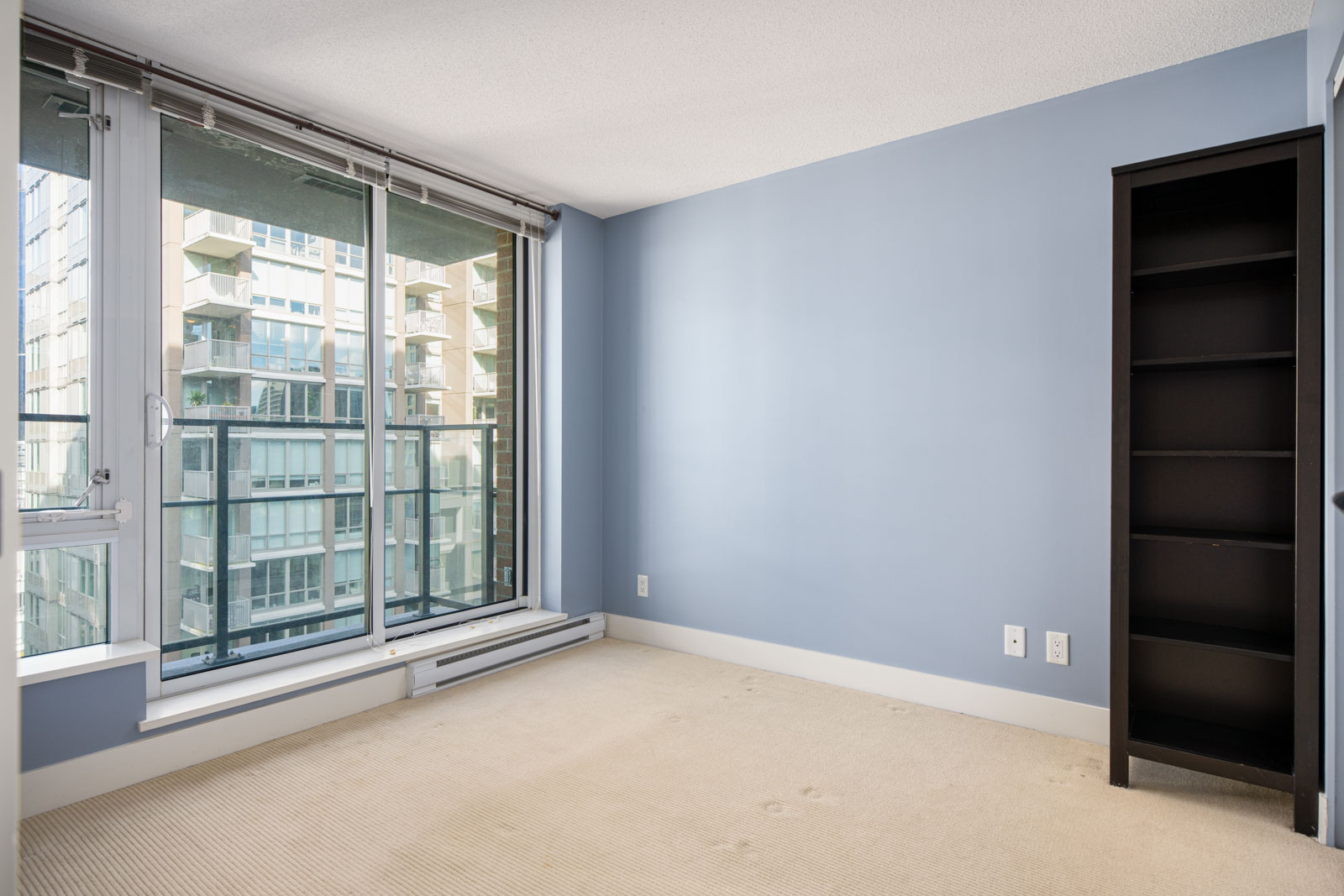 Empty room with light blue walls, beige carpet, large window and glass door leading to a balcony, black shelving unit, and view of adjacent building.