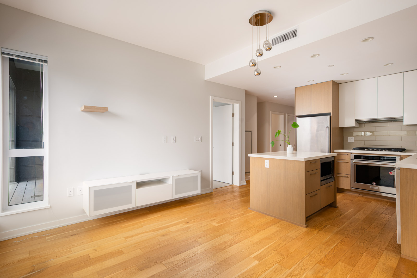 Modern, unfurnished kitchen and living area with wood floors, light wood cabinets, white countertops, and pendant lights; a window and built-in wall unit are visible.