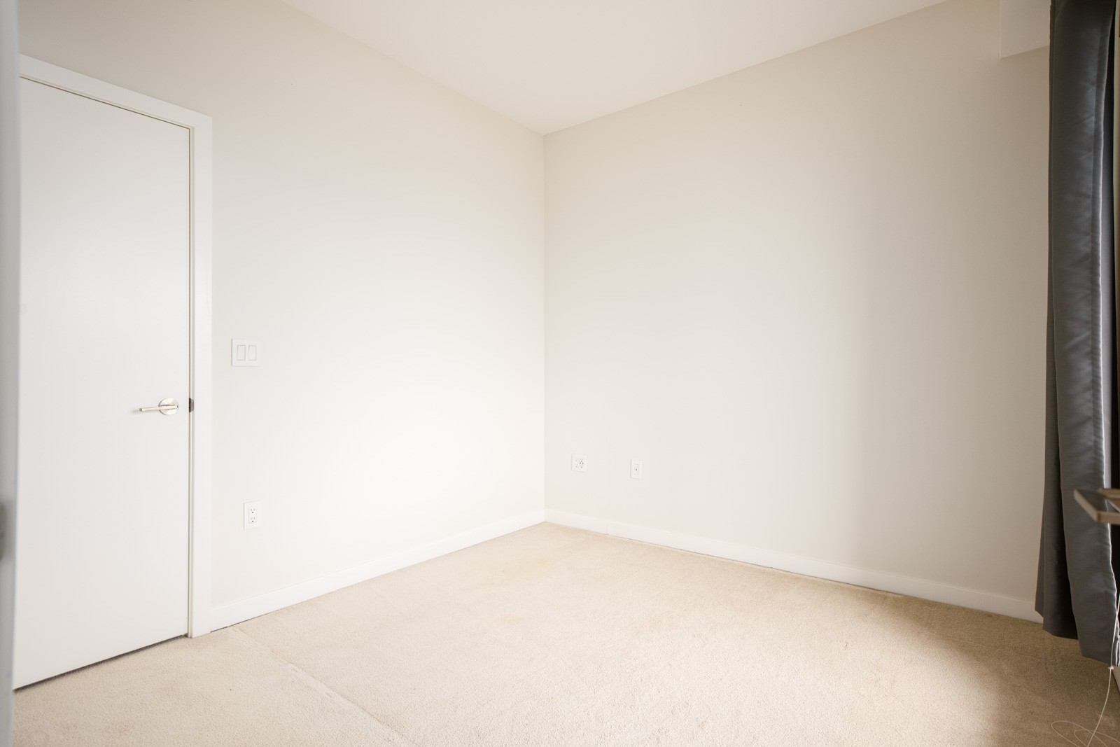 An empty corner of a room with beige carpet, white walls, a closed white door, electrical outlets, and a window with dark curtains on the right.