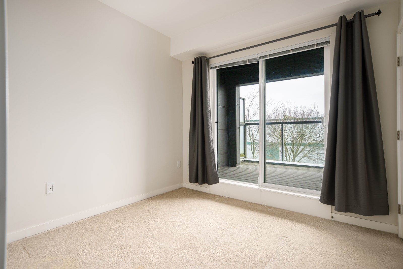 Empty room with beige carpet, white walls, and a glass sliding door leading to a balcony with dark curtains on each side; view of trees and water outside.