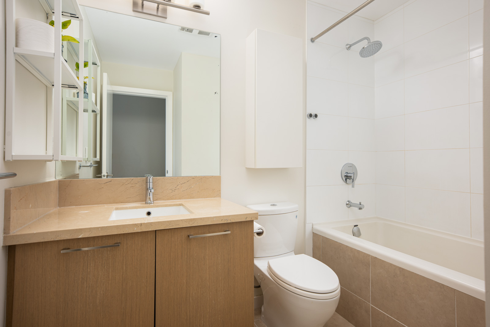 Modern bathroom with a wooden vanity, sink, toilet, white tile bathtub-shower combo, wall mirror, and shelving unit with towels and plants.
