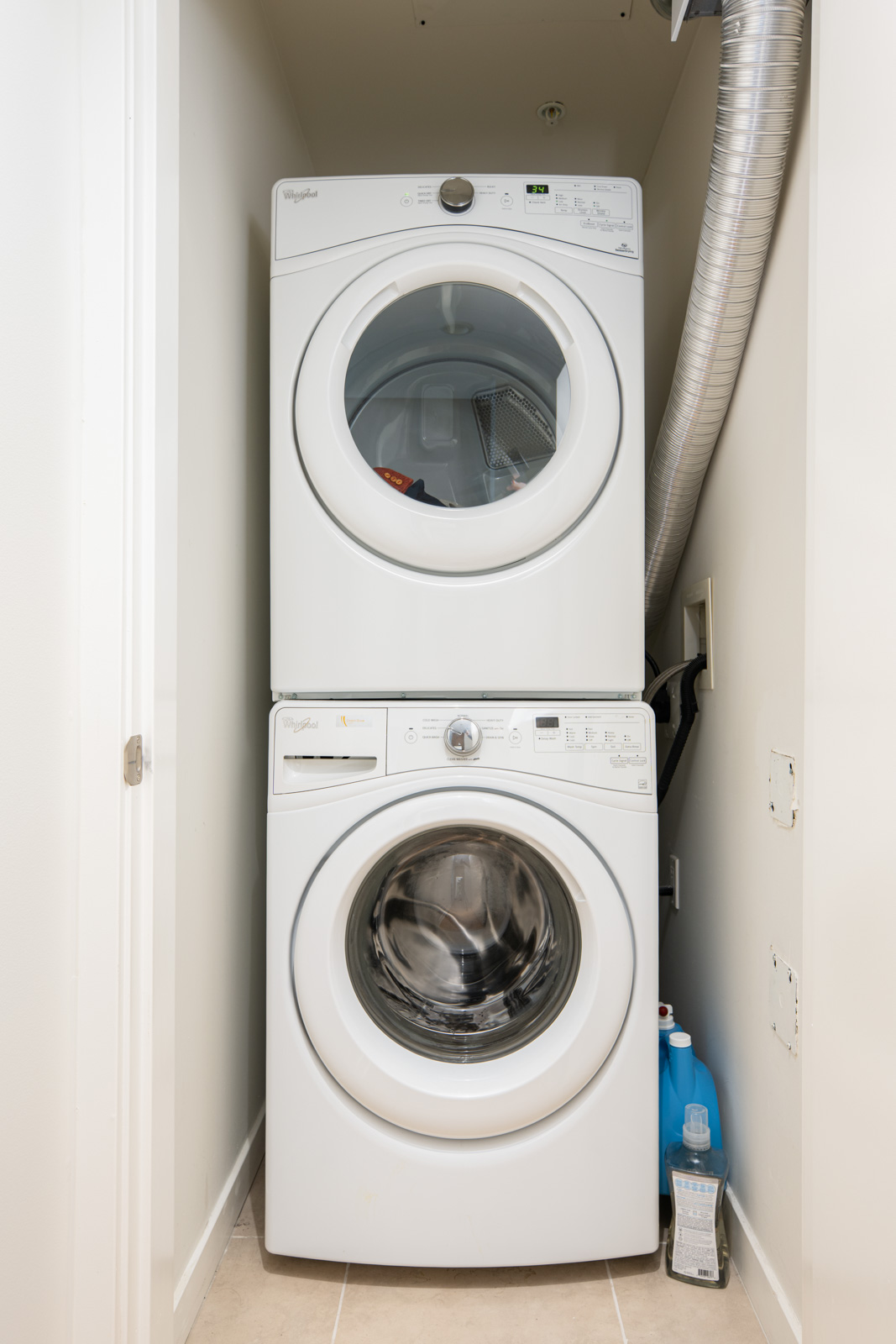 A stacked washer and dryer unit is installed in a small laundry closet, with detergent bottles stored on the floor to the right.