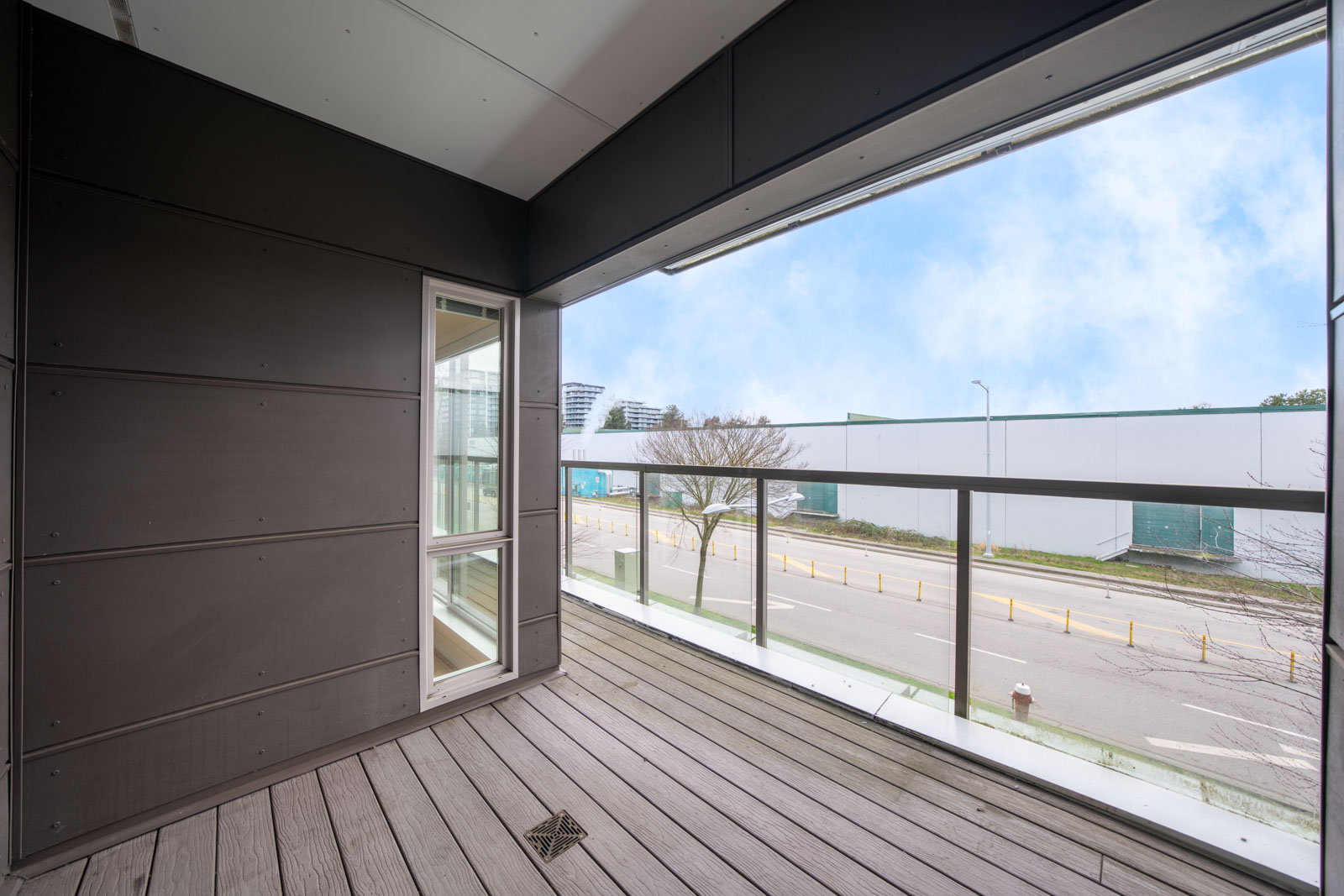Empty balcony with wood flooring, glass railing, and a view of a street, tree, and industrial buildings under a partly cloudy sky.