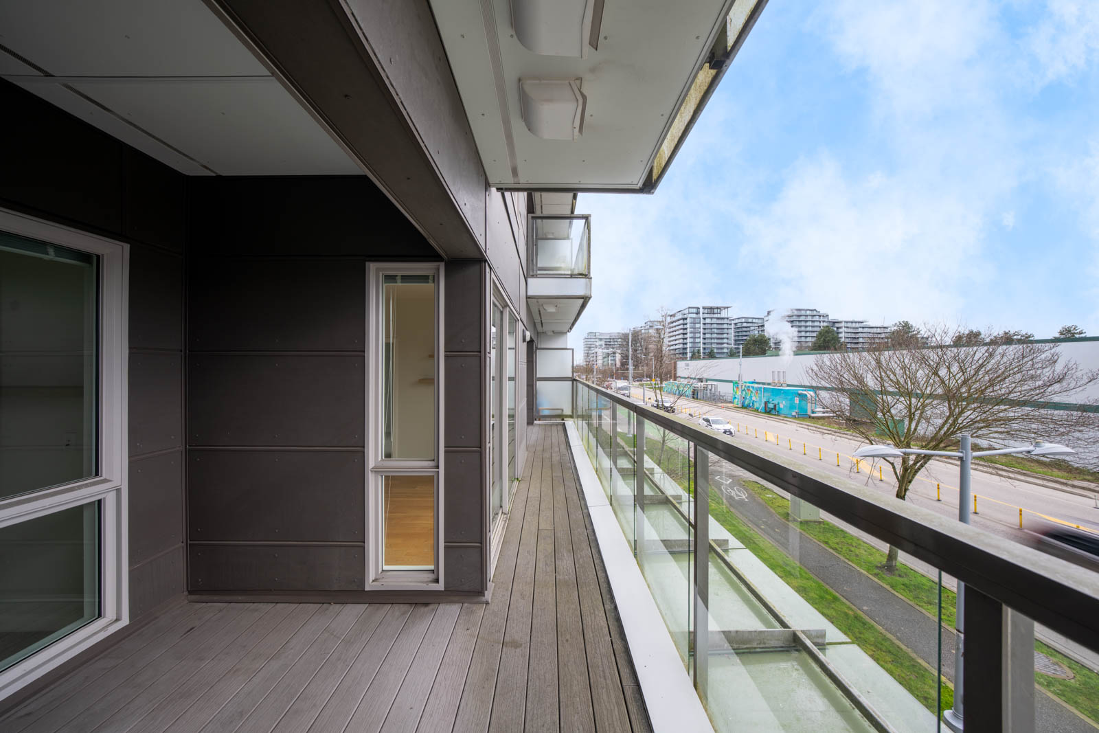 Modern apartment balcony with glass railing overlooking a street, nearby buildings, and trees under a partly cloudy sky.