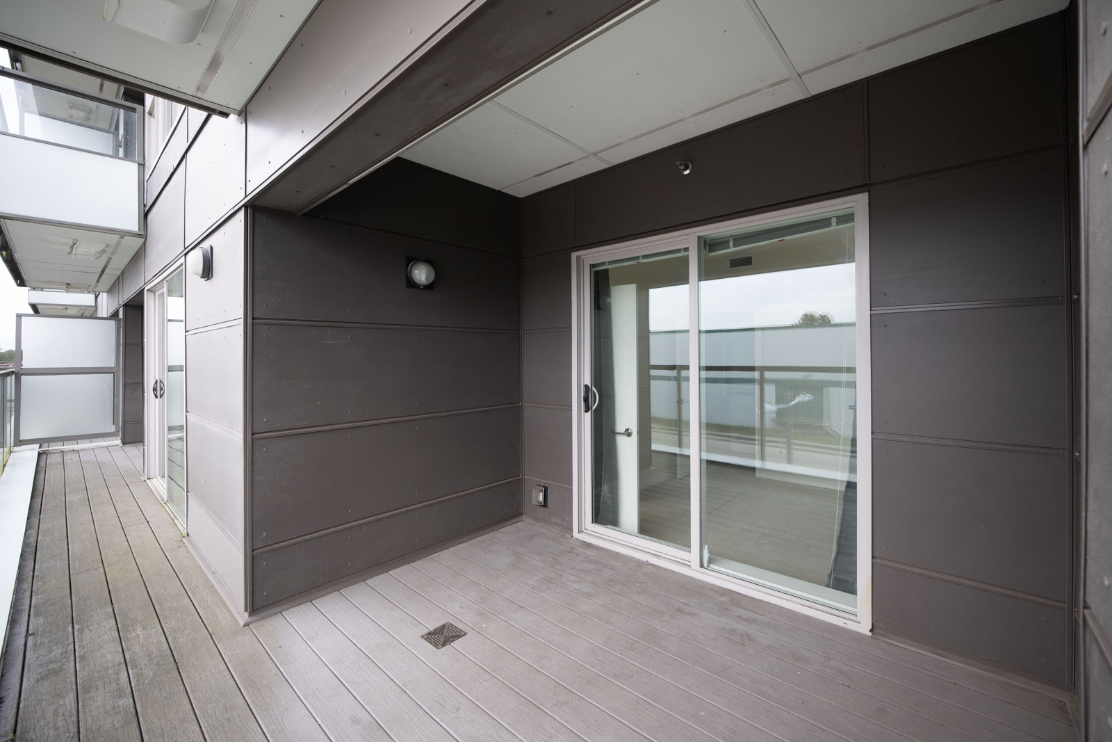 A modern, empty balcony with wood flooring, dark panel walls, ceiling lighting, and a sliding glass door leading inside.