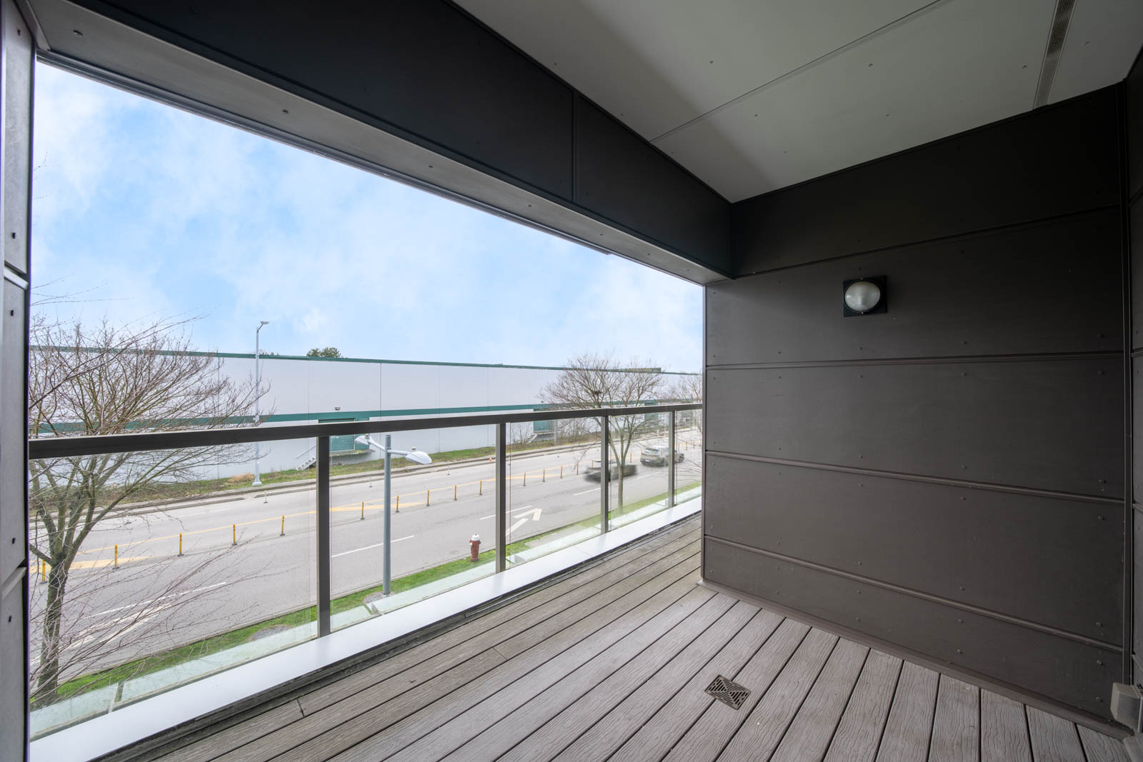 A modern balcony with wooden flooring and glass railing overlooks a street with trees, industrial buildings, and a cloudy sky.