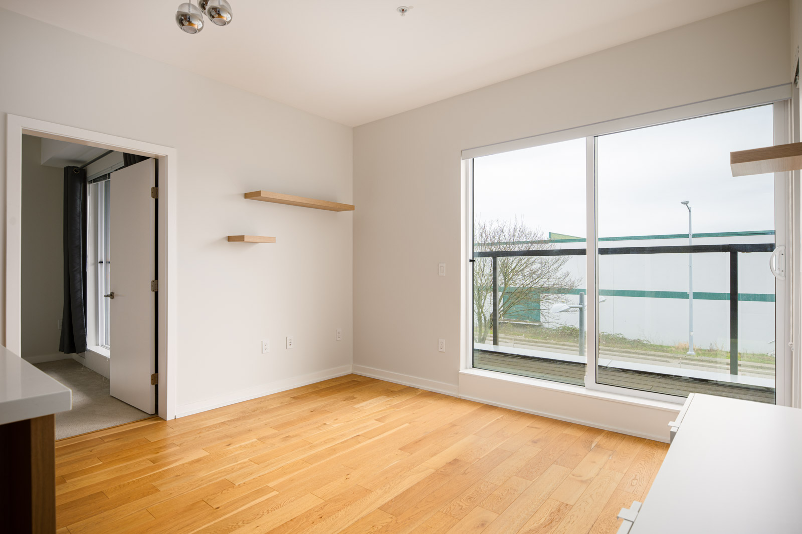 Empty room with light wood flooring, white walls, large window, two floating shelves, and an open door leading to another room.