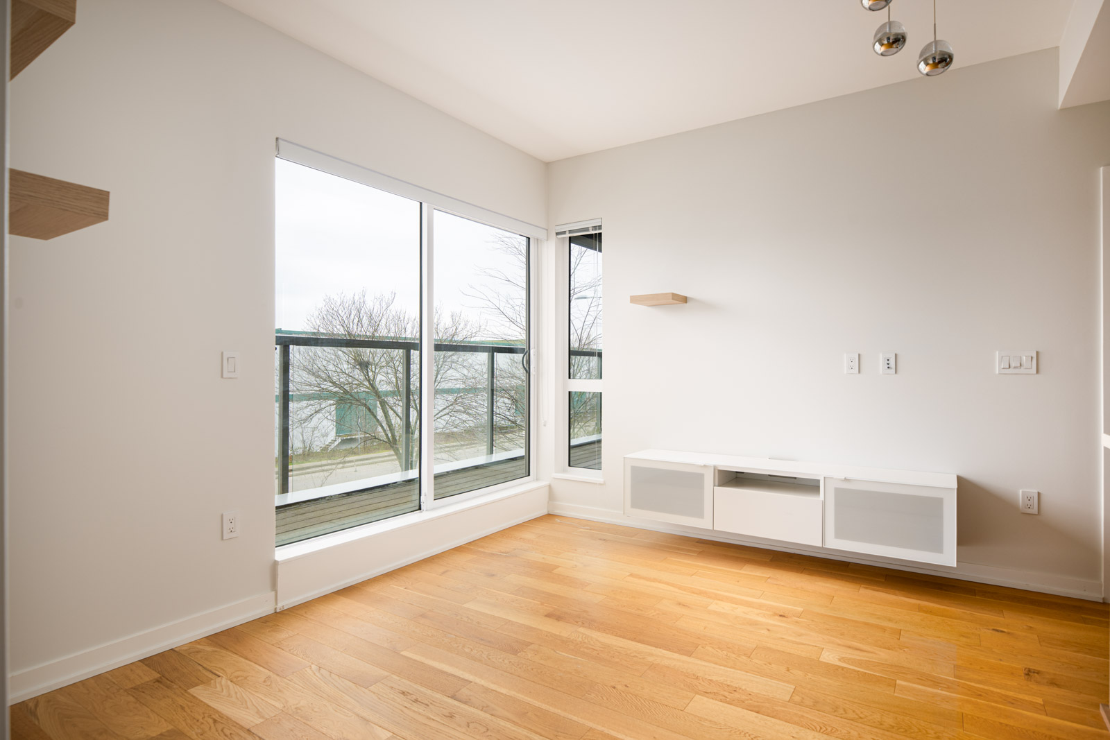 A bright, empty room with light wood flooring, large windows overlooking trees and water, white walls, and a floating media cabinet.