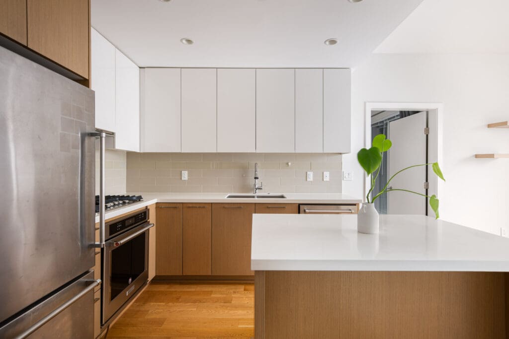 Modern kitchen with wooden cabinets, white countertops, stainless steel appliances, and a potted plant on the island. White tile backsplash and minimal decor.