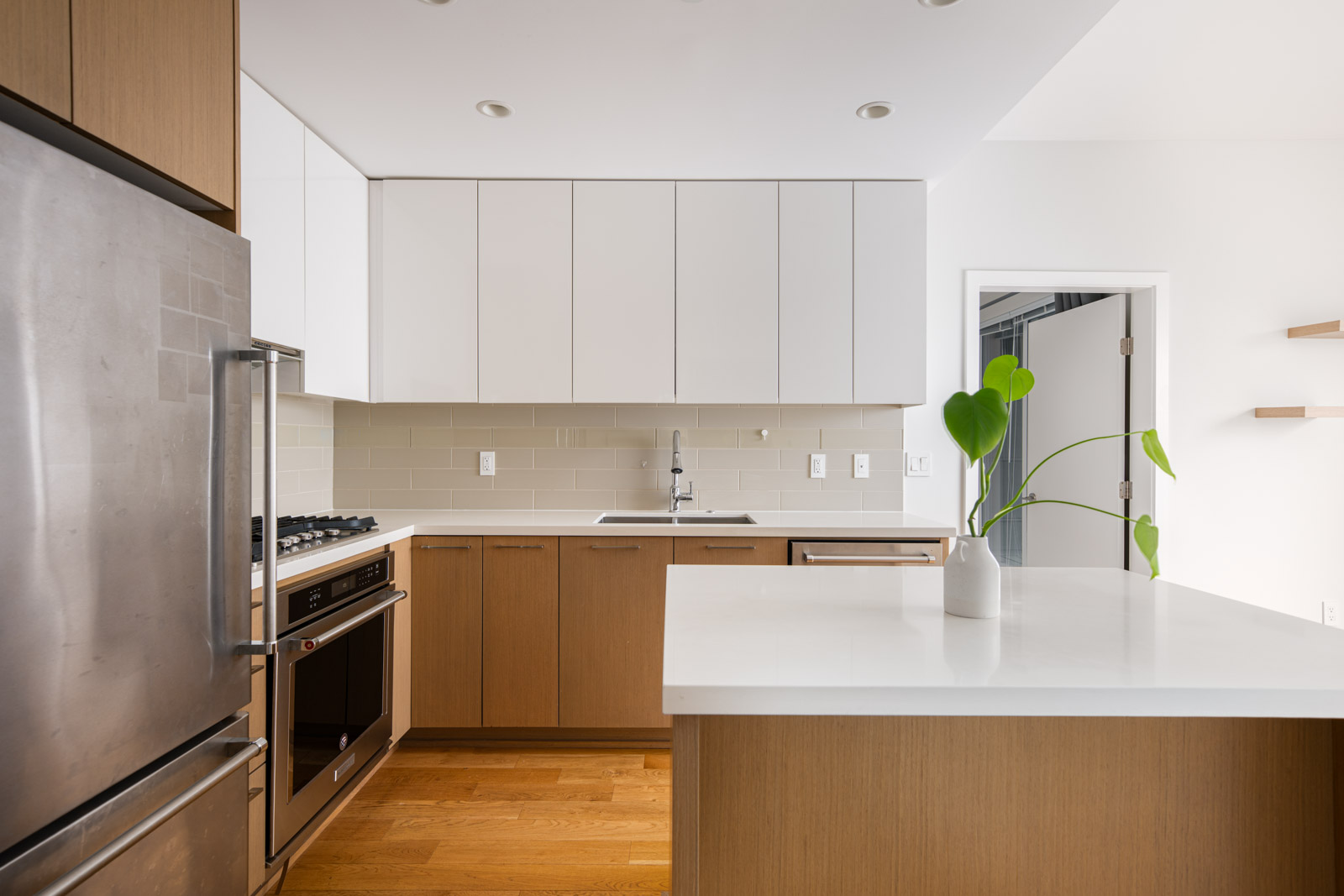 Modern kitchen with wooden cabinets, white countertops, stainless steel appliances, and a potted plant on the island. White tile backsplash and minimal decor.