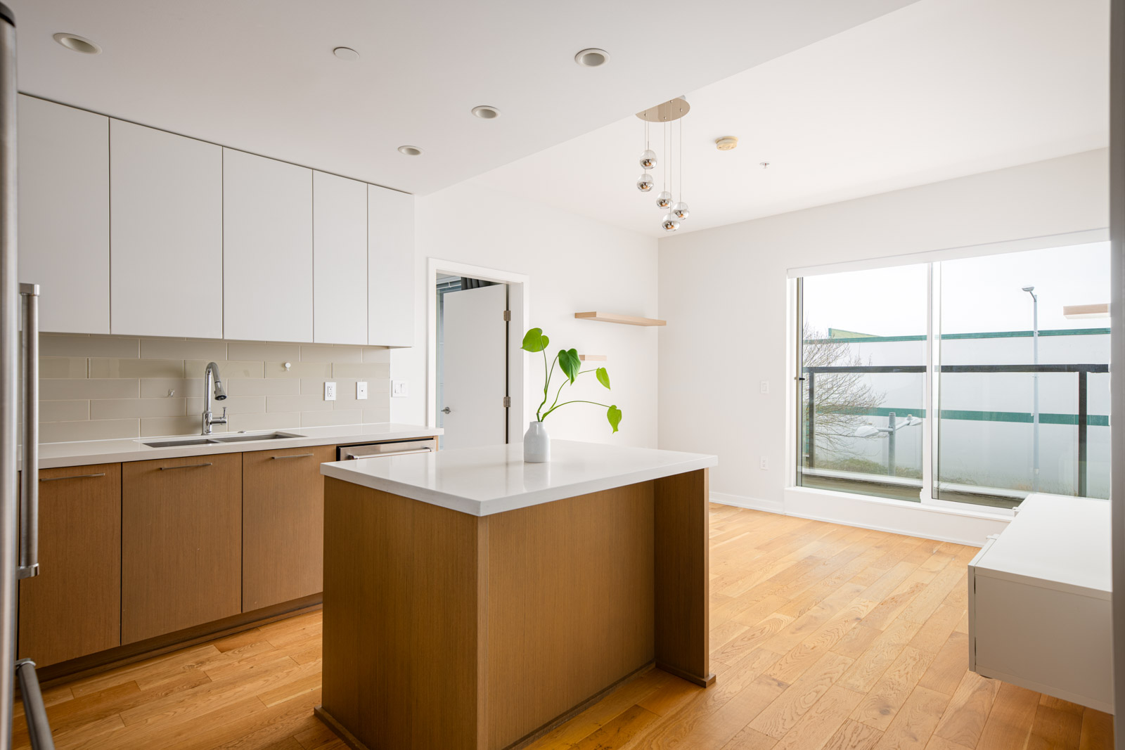 Modern kitchen with wood floors, white cabinetry, a central island, large window, and a potted plant on the island. The space is bright and minimalistic.