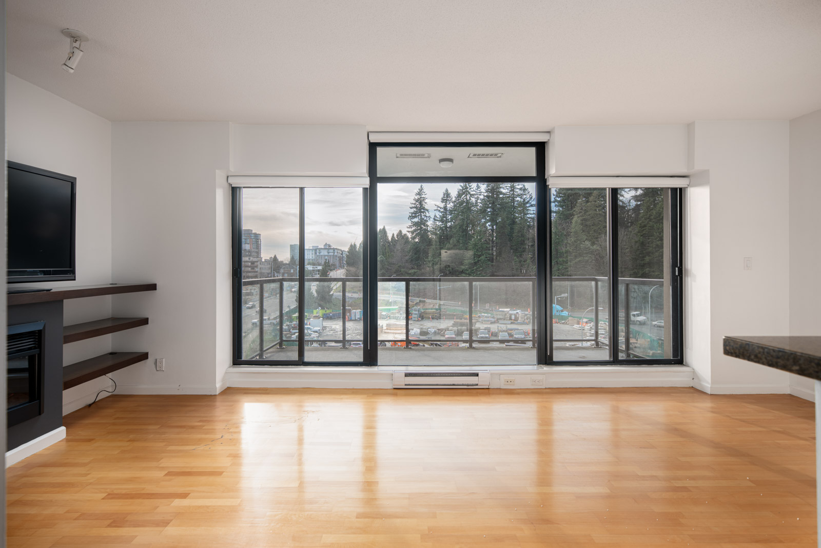 Empty living room with light wood floors, large windows, and a balcony overlooking a construction site and trees outside.