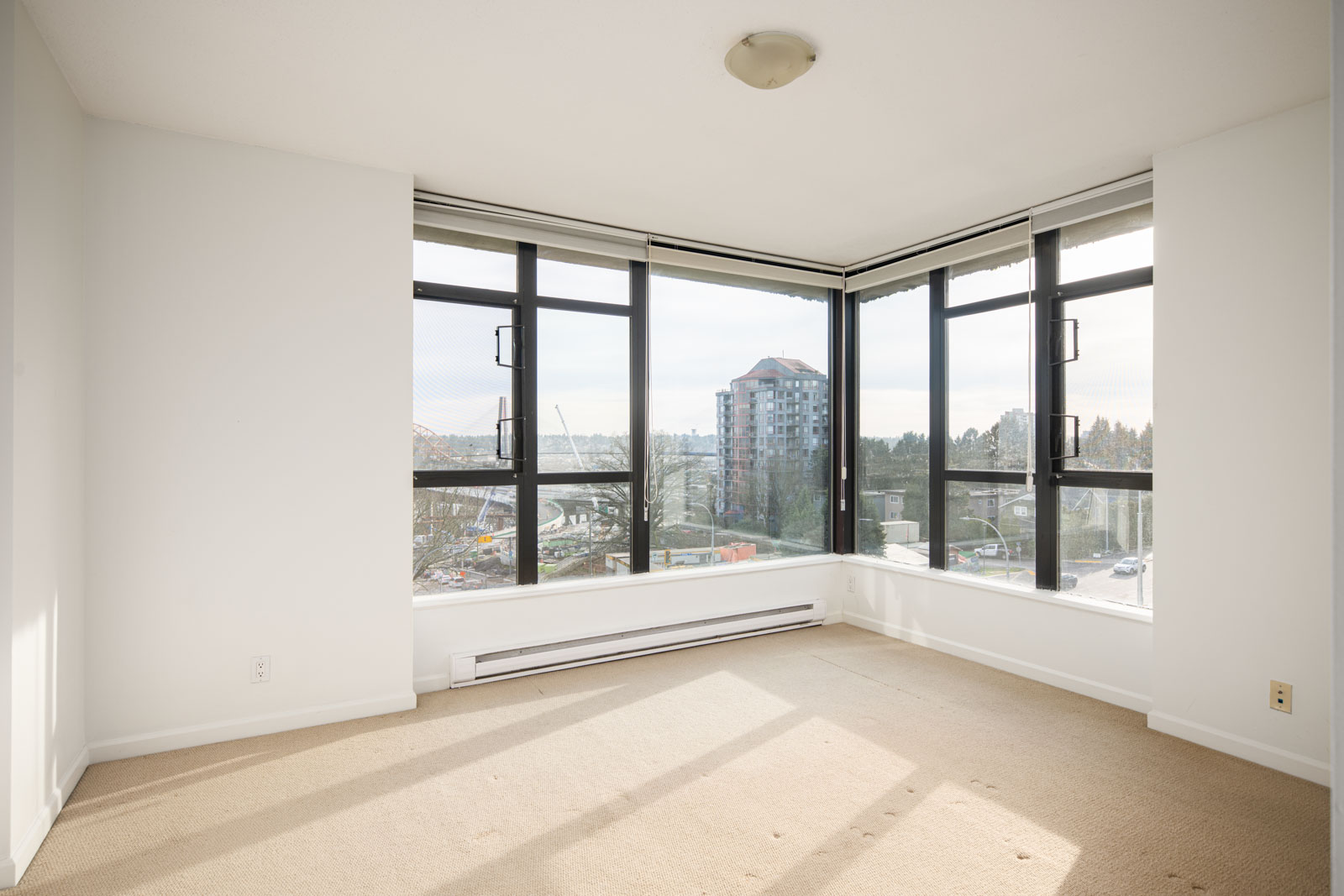 Empty corner room with large windows, beige carpet, and a view of a cityscape including a tall building and distant trees. Natural light fills the space.