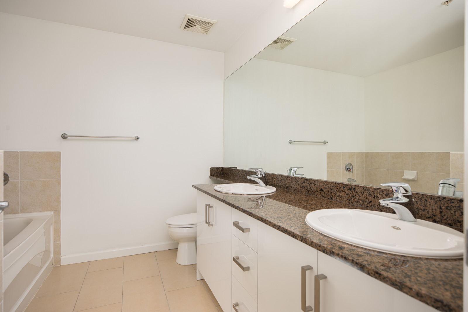 Modern bathroom with double sinks, granite countertop, large mirror, white cabinets, a bathtub, and beige tile flooring.
