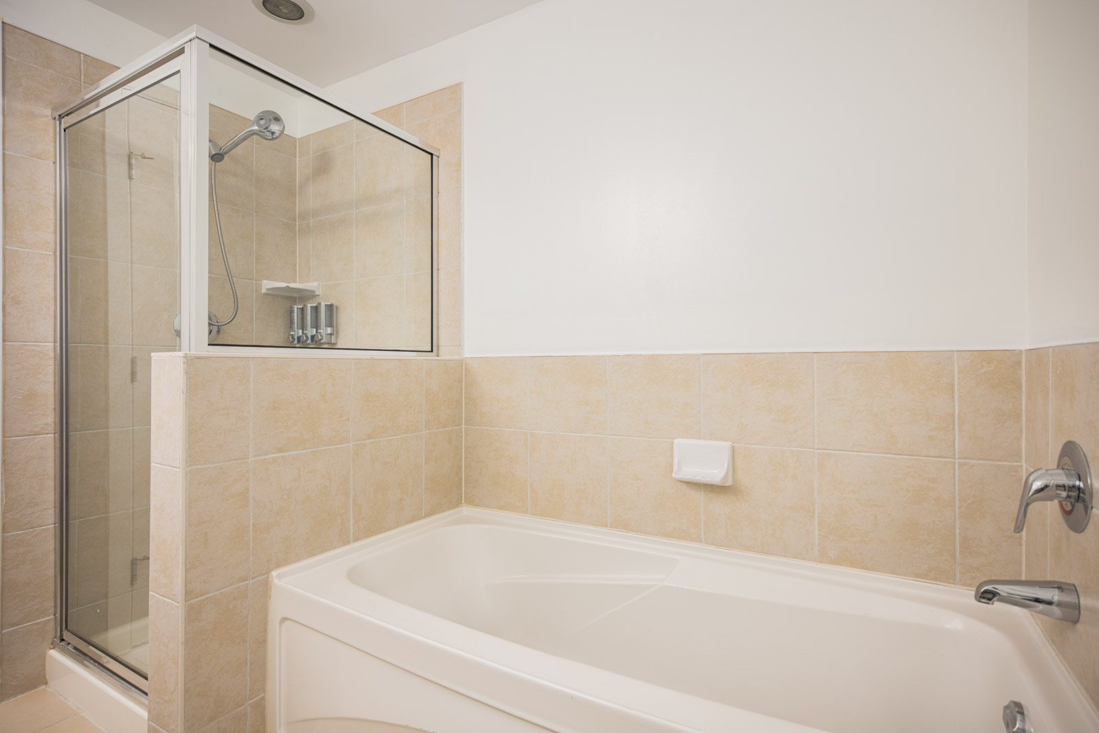 A bathroom with a glass-enclosed shower and a rectangular bathtub, both surrounded by beige tile walls.