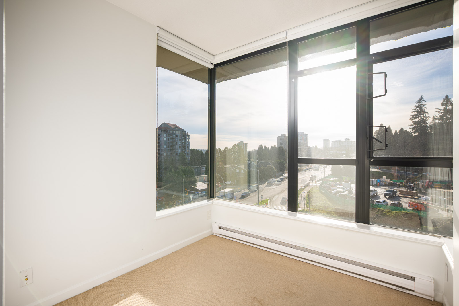 Empty corner of a room with large floor-to-ceiling windows, beige carpet, white walls, and a view of buildings and trees outside on a sunny day.