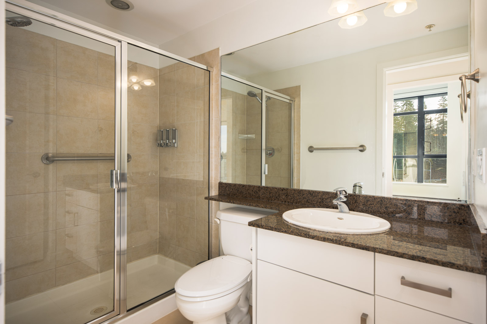 Modern bathroom with a glass-enclosed shower, granite countertop, white sink and cabinets, toilet, and a window letting in natural light.
