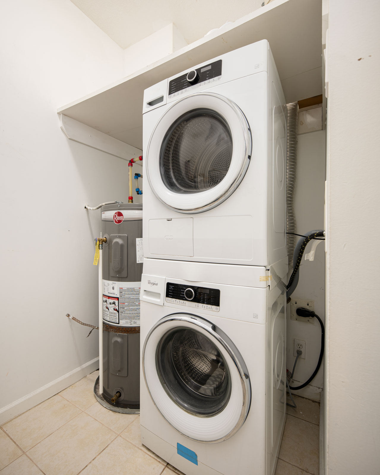 Stacked washer and dryer units next to a water heater in a small laundry room with tiled floor and white walls.