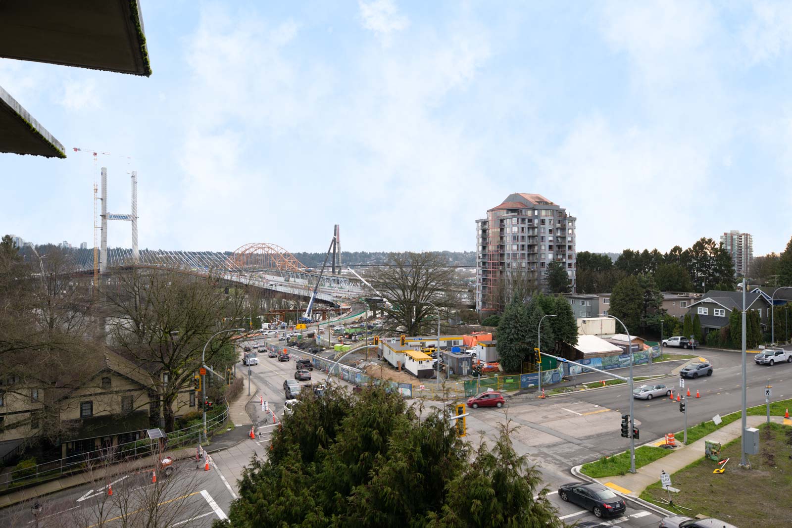 A cityscape shows a bridge under construction, an apartment building, houses, and traffic at an intersection with roadwork and construction equipment.