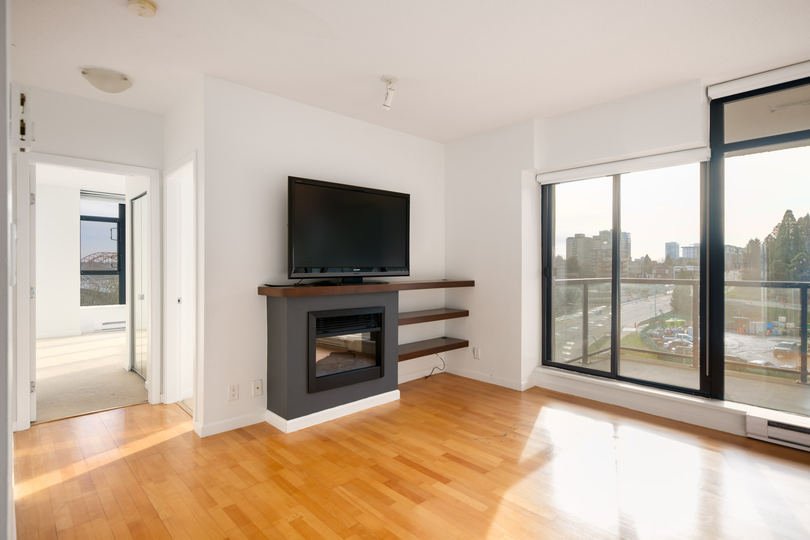 Bright living room with hardwood floors, wall-mounted TV above a fireplace, large window, sliding glass door to a balcony, and an open door leading to a bedroom.