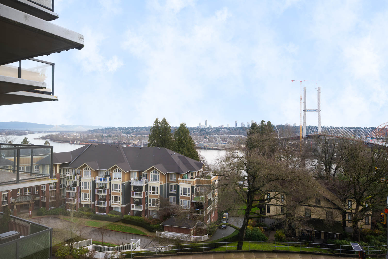 View from a balcony overlooking apartment buildings, a river, trees, and a distant bridge under a partly cloudy sky.
